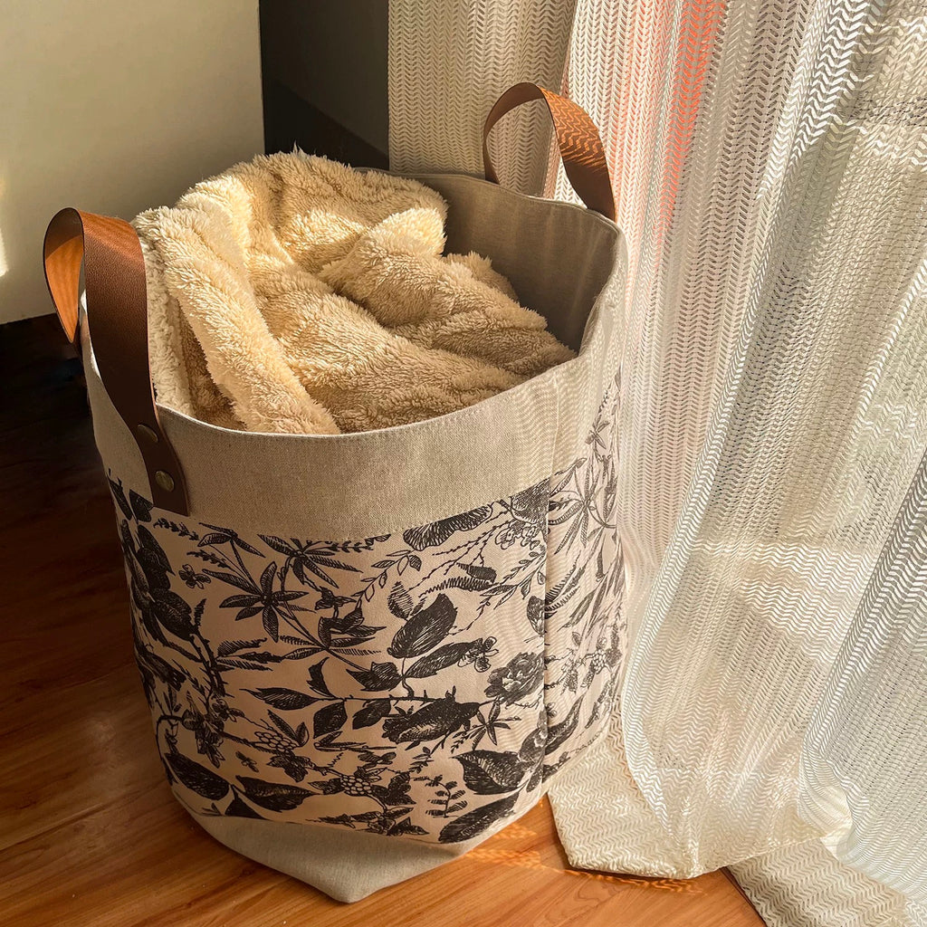 Floral-patterned laundry basket with a beige blanket inside, placed on a wooden floor.