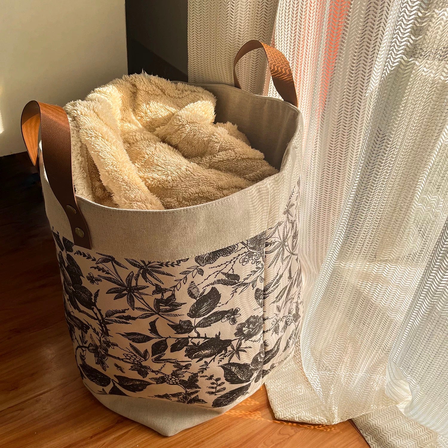 Floral-patterned laundry basket with a beige blanket inside, placed on a wooden floor.