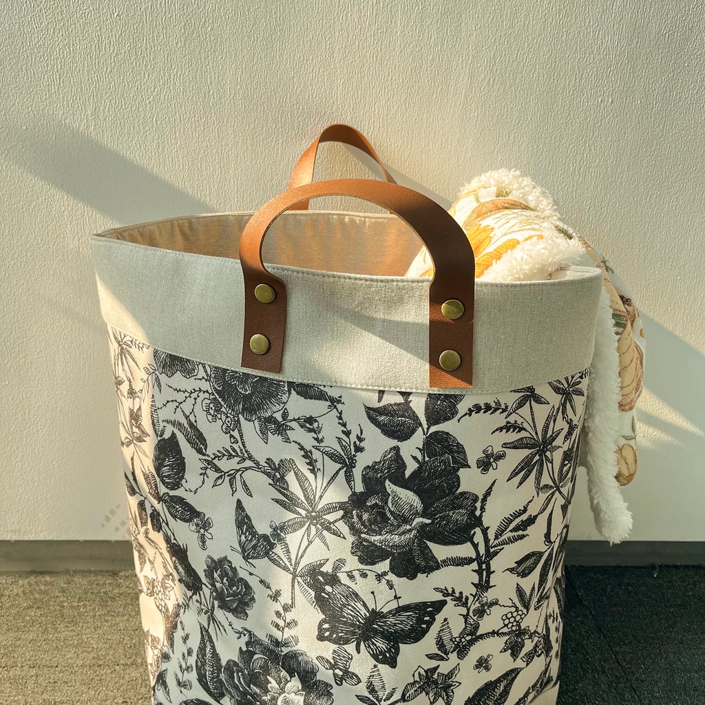 Tote bag with floral pattern and brown leather handles against a plain wall.